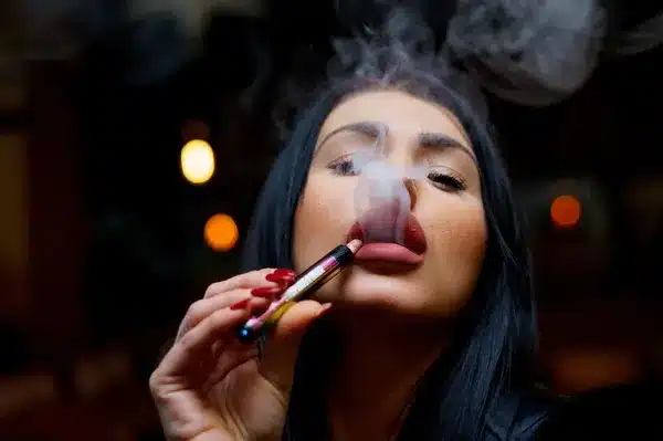 Una mujer sostiene un dispositivo mientras exhala vapor; la escena puede asociarse al uso de vapes, precio variable según modelo.